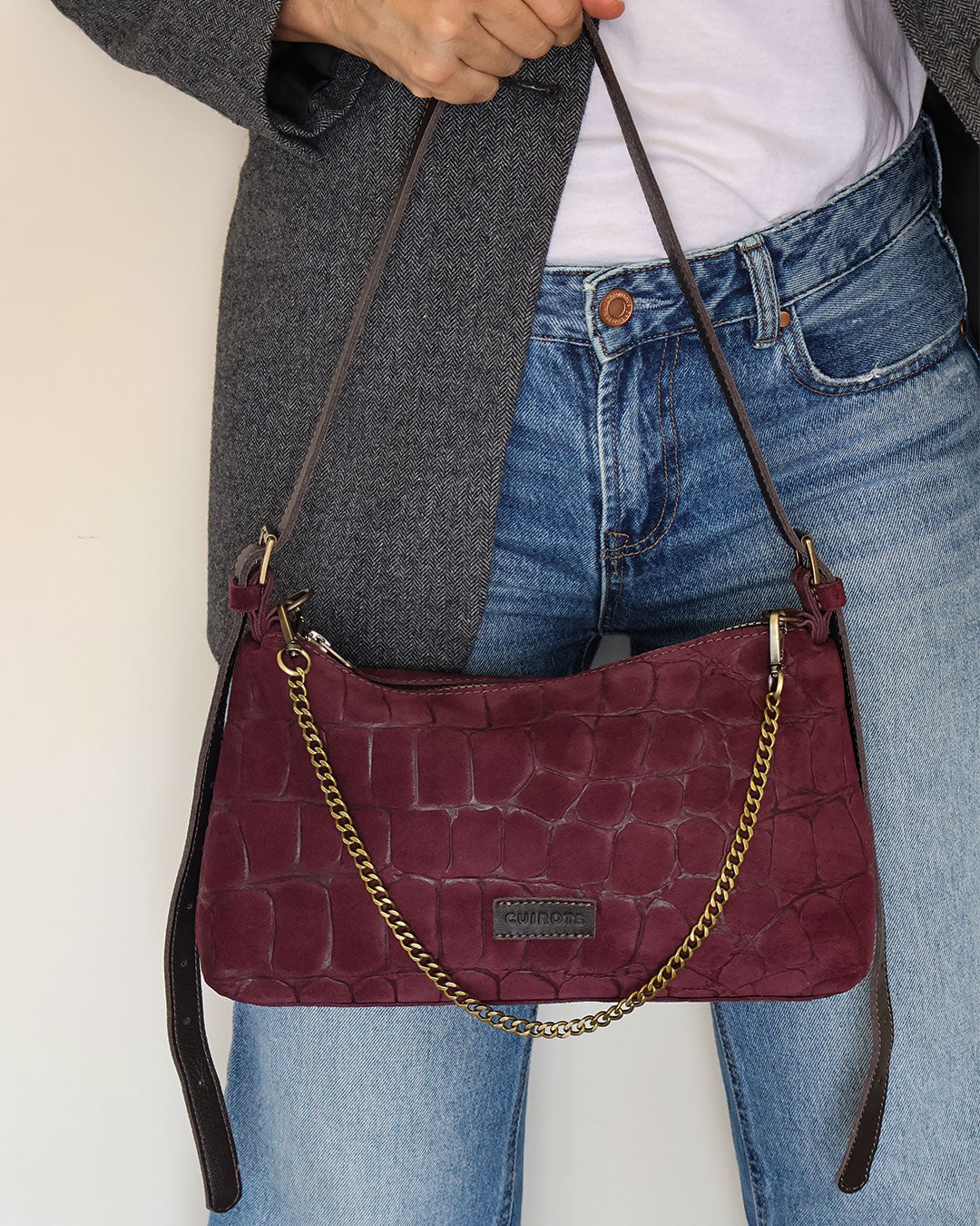 Bolso de hombro y bandolera Coco de serraje en color burdeos, con asa de cadena, sujetado por el asa de piel sobre unos vaqueros