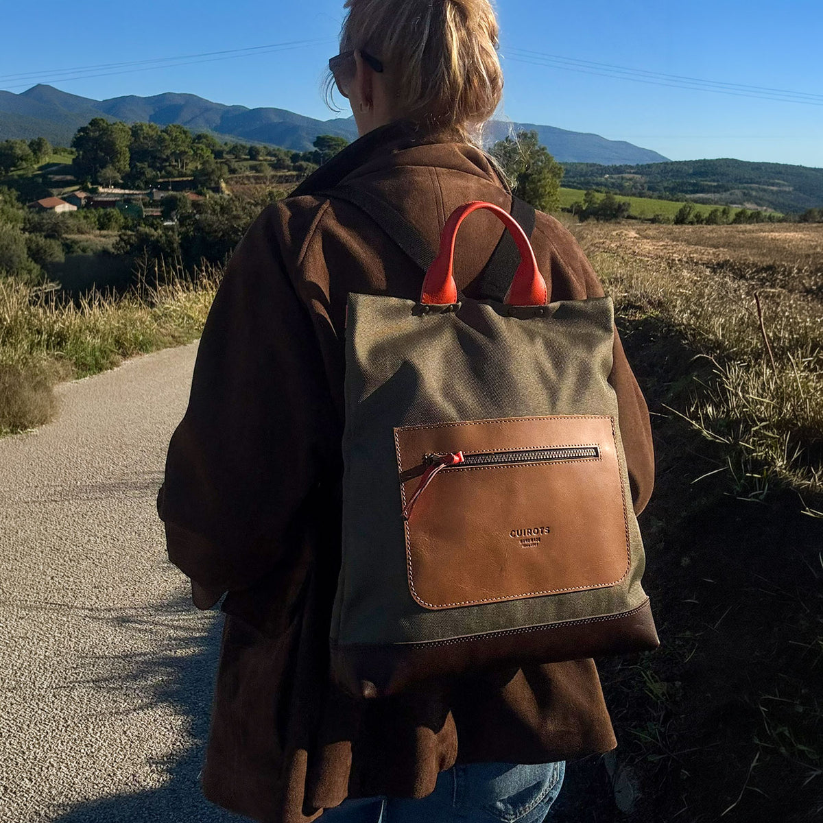 Mujer de espaldas con la mochila antirrobo Trend kaki de lona y piel, en un camino de montaña